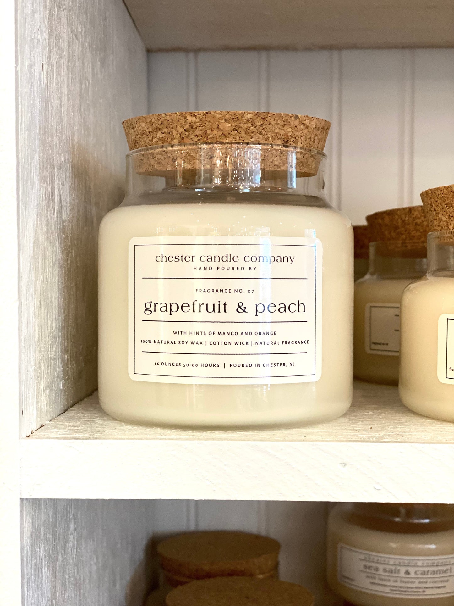 Natural Soy Wax Candle in a Clear Glass Apothecary-Style Jar and a Cork Lid. White Label on the Jar Reads "chester candle company. Fragrance No. 07. Grapefruit & peach with hints of mango and orange. 100% Natural Soy Wax, Cotton Wick, Natural Fragrance. 16 ounces 50-60 hours. Poured in Chester, NJ”