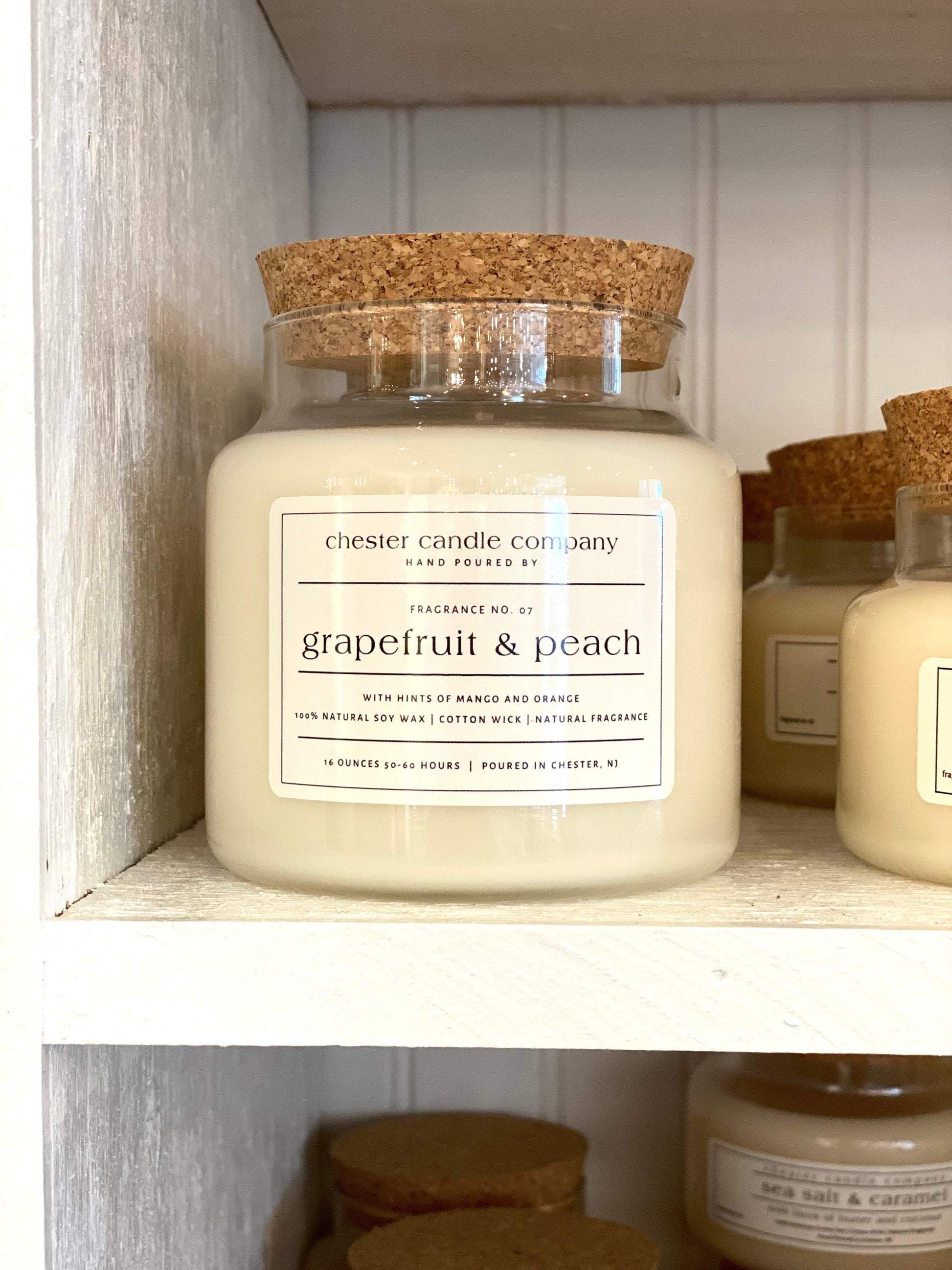 Natural Soy Wax Candle in a Clear Glass Apothecary-Style Jar and a Cork Lid. White Label on the Jar Reads "chester candle company. Fragrance No. 07. Grapefruit & peach with hints of mango and orange. 100% Natural Soy Wax, Cotton Wick, Natural Fragrance. 16 ounces 50-60 hours. Poured in Chester, NJ”